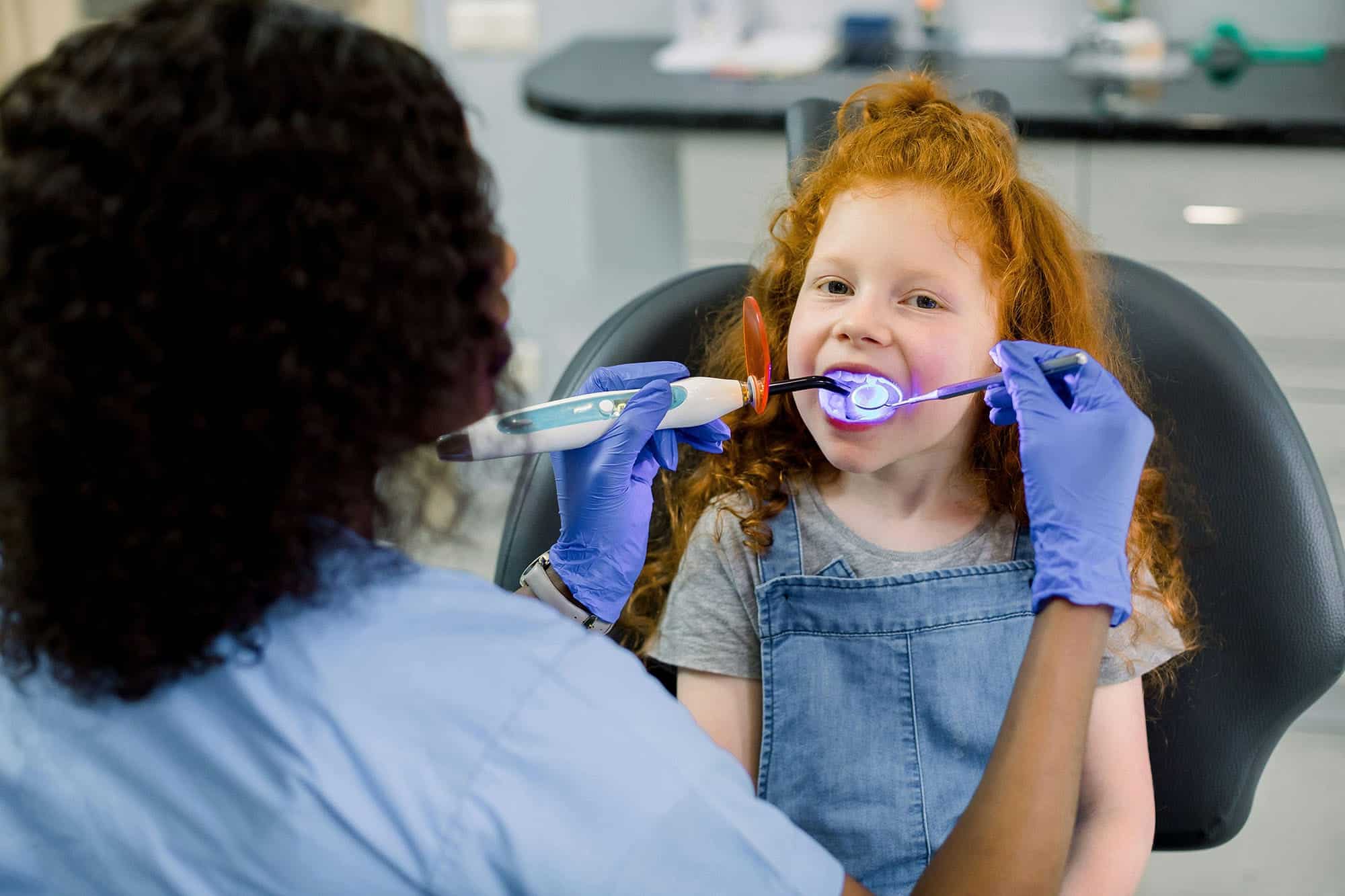 A dentist wearing gloves works with a smiling young girl in a dental chair during a checkup.