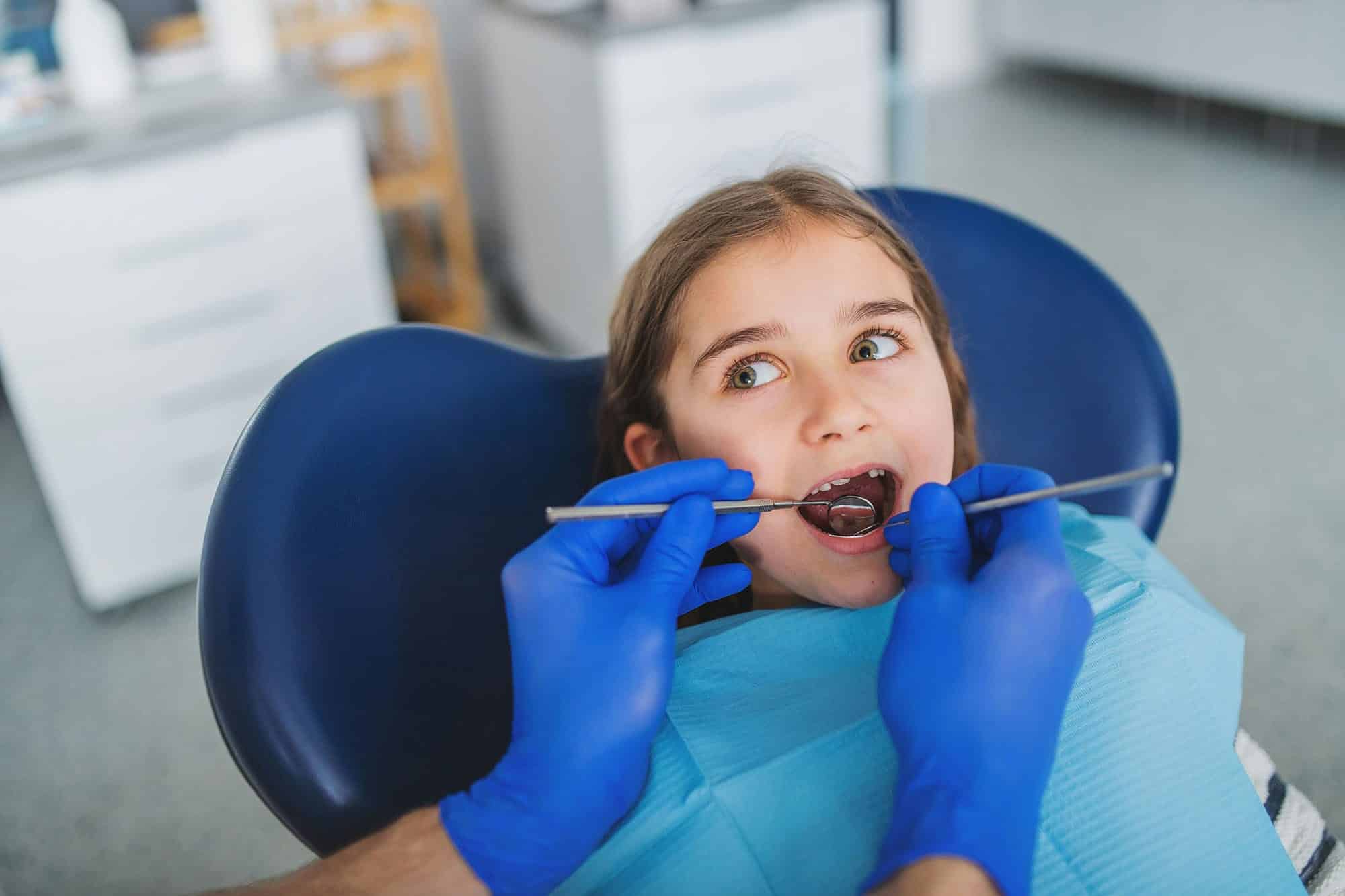 A dentist wearing blue gloves examines a young girl's teeth in a dental chair.