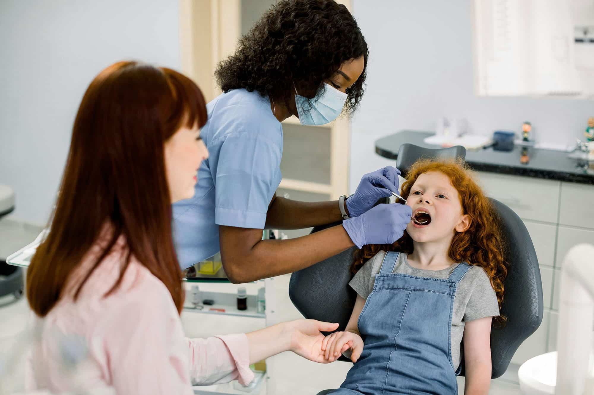 A dentist wearing a mask and gloves provides children's dental services, examining a young girl’s mouth in a dental chair while a caregiver sits nearby.