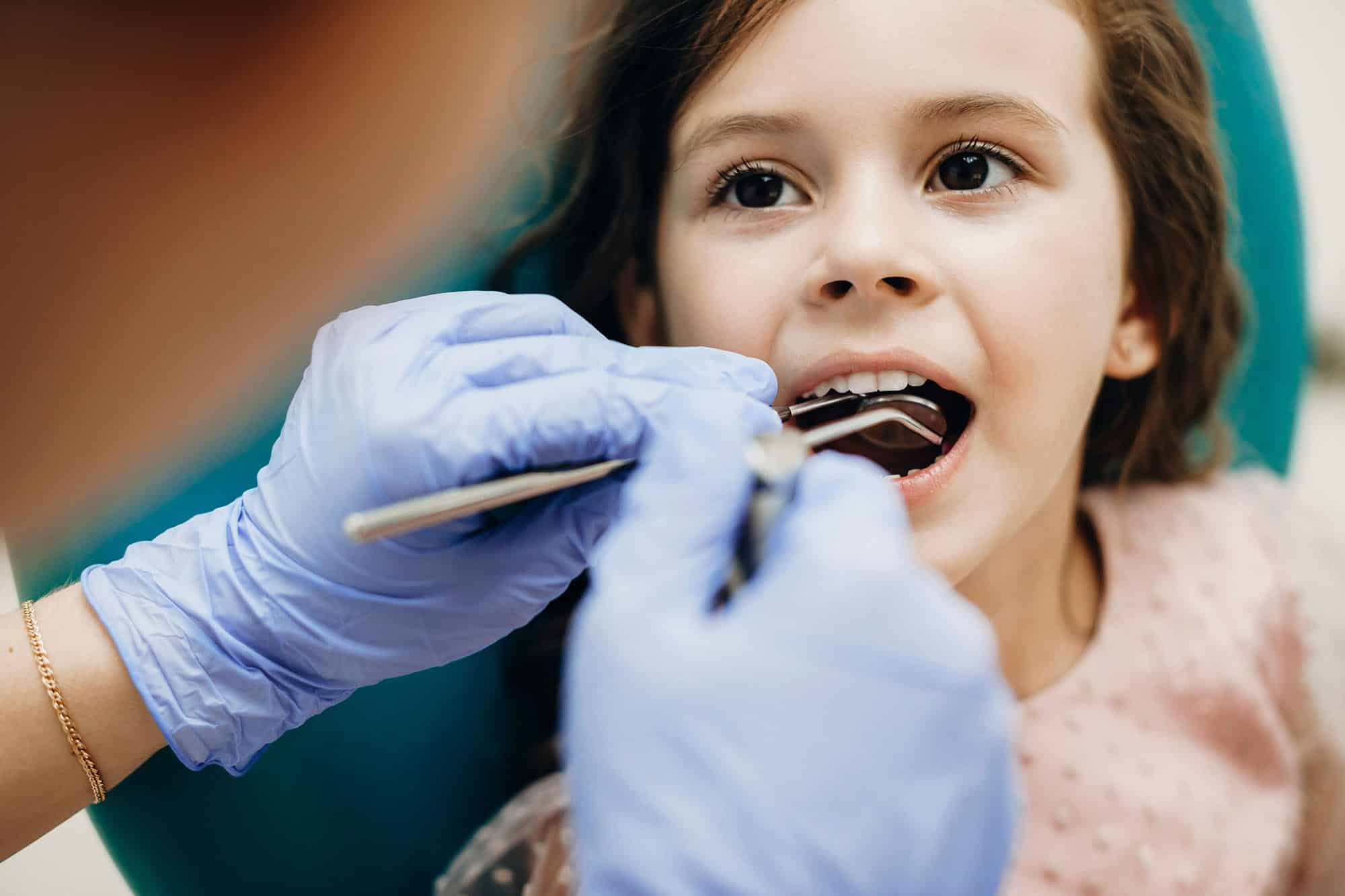 A young girl sits in a dental chair while a dentist in blue gloves examines her mouth with dental tools.