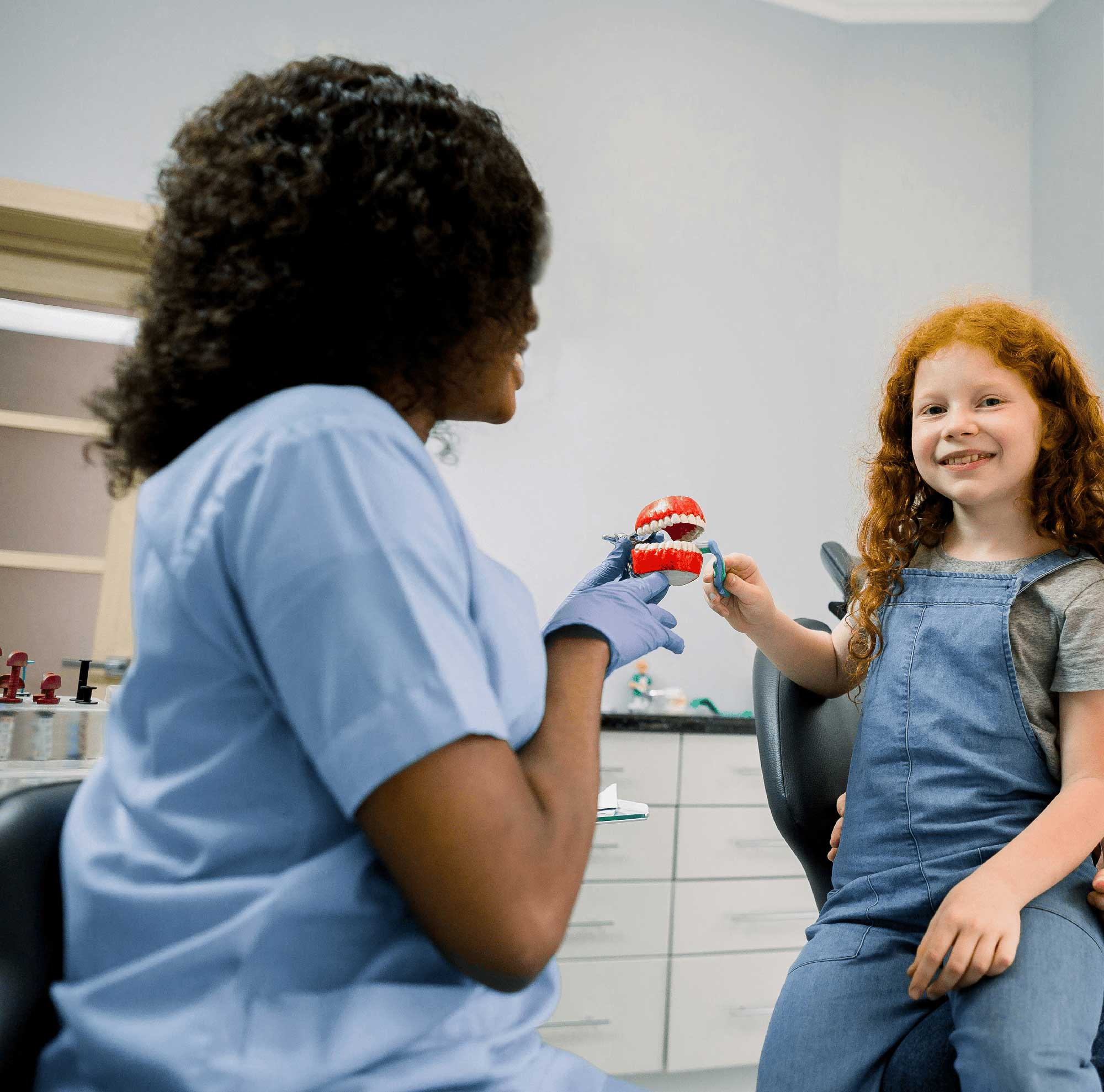 A healthcare professional in blue scrubs and gloves hands a red and blue toy to a smiling red-haired girl in a pediatric dental clinic.