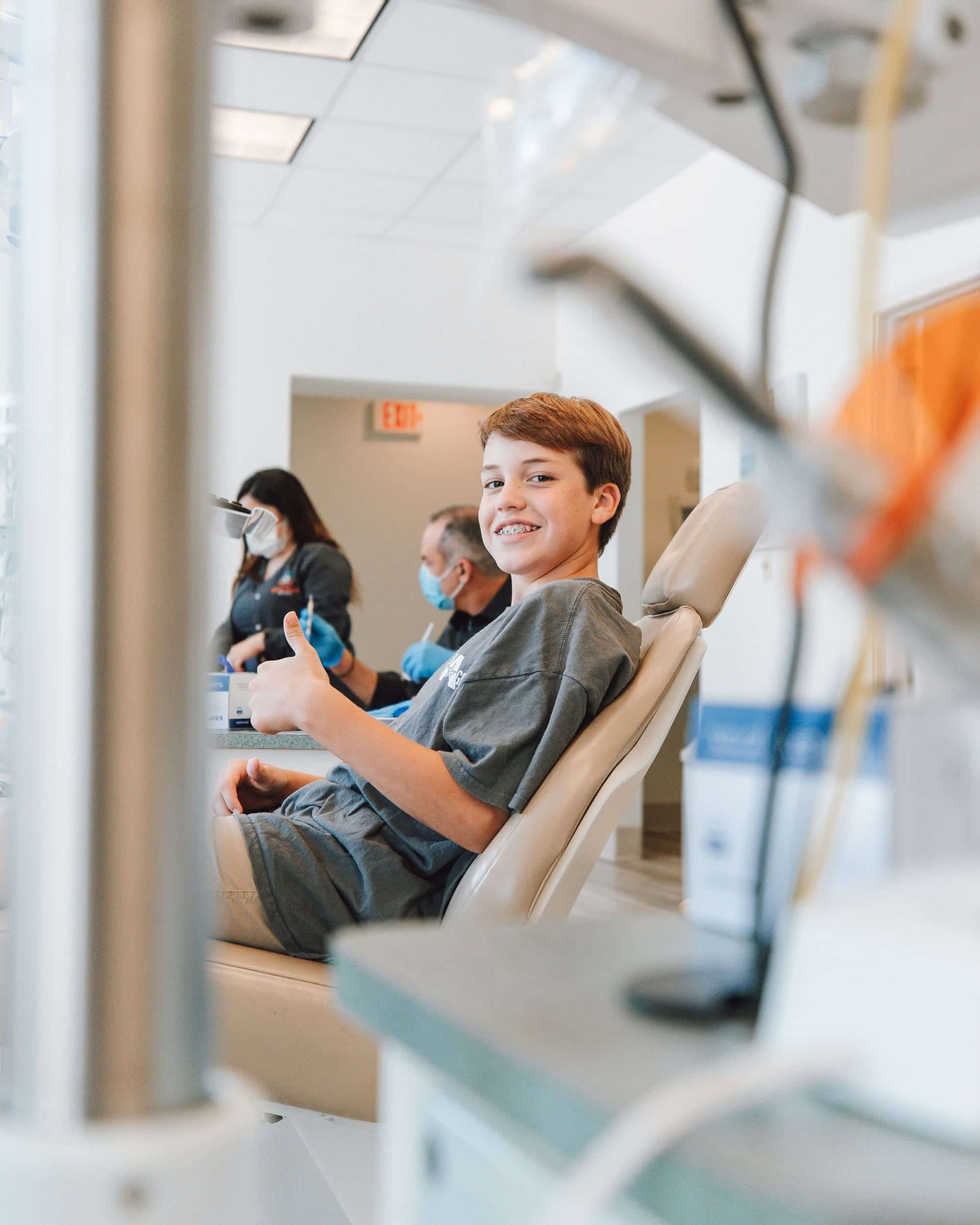 A smiling patient sits in a dental chair in a clinic, with masked staff in the background.